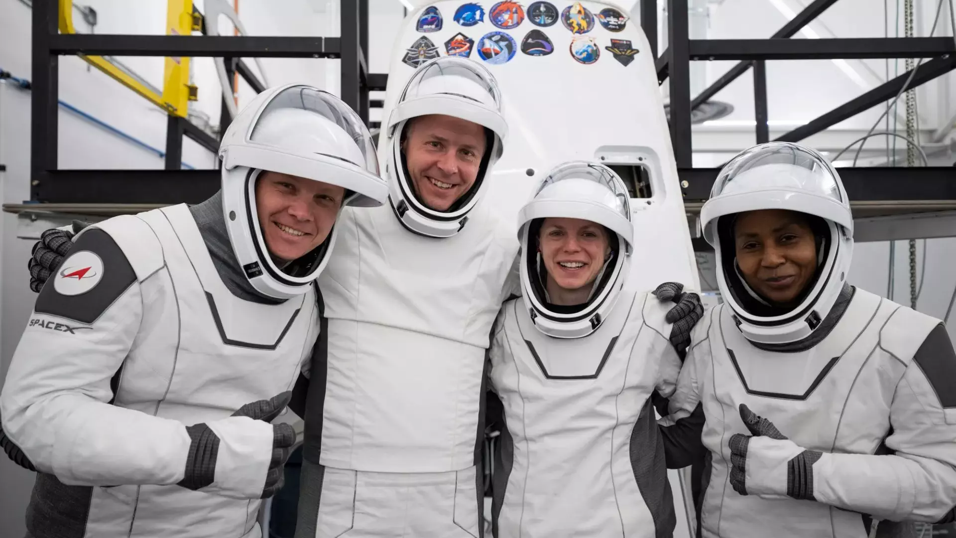 NASA Space Technology four people in white spacesuits with the visors up pose and smile in front of a white cone-shaped spacecraft