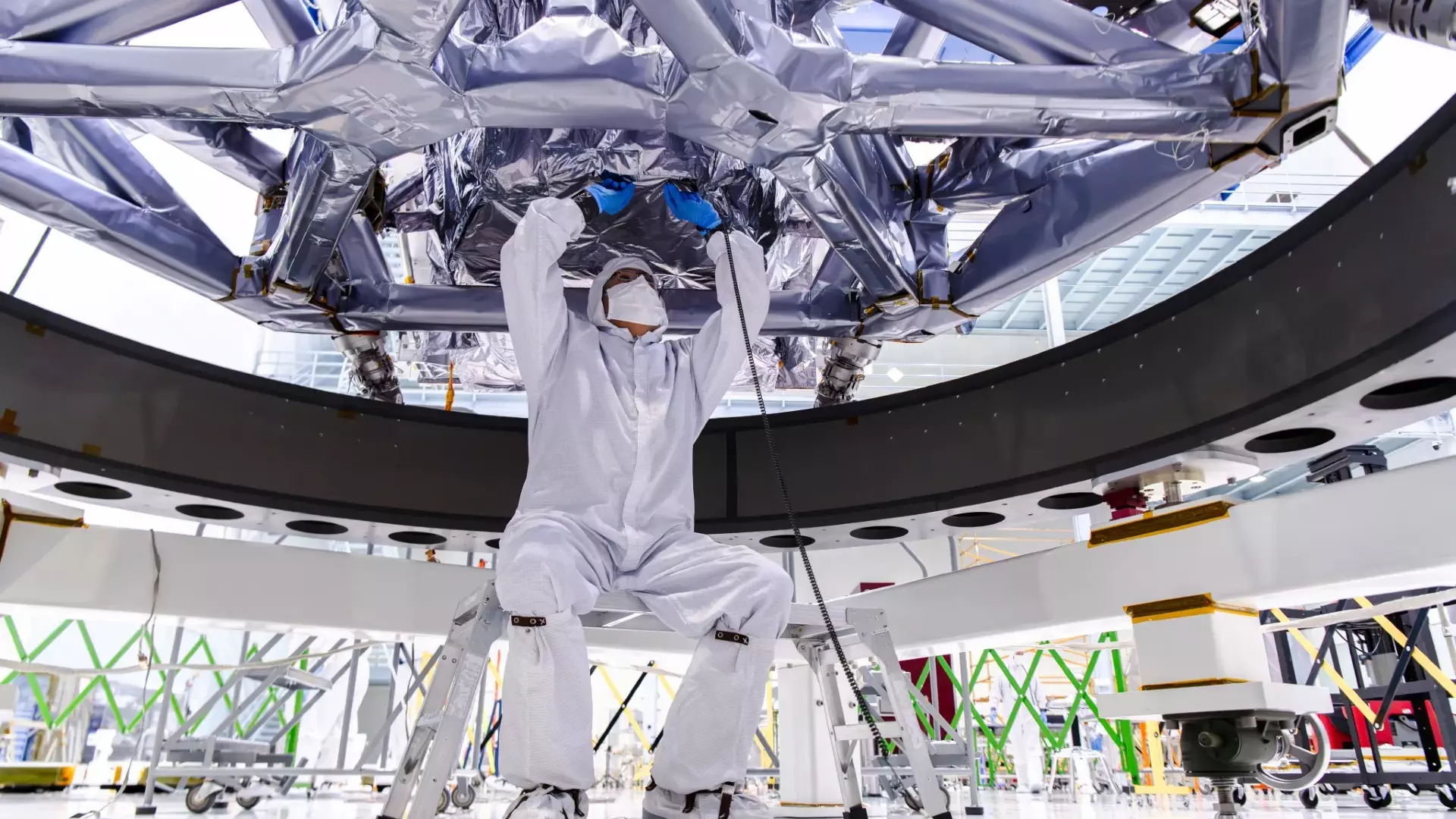 NASA Space Technology scientists in white clean suits move a metallic honeycomb-like structure onto a rectangular piece of electronics covered in wires and components