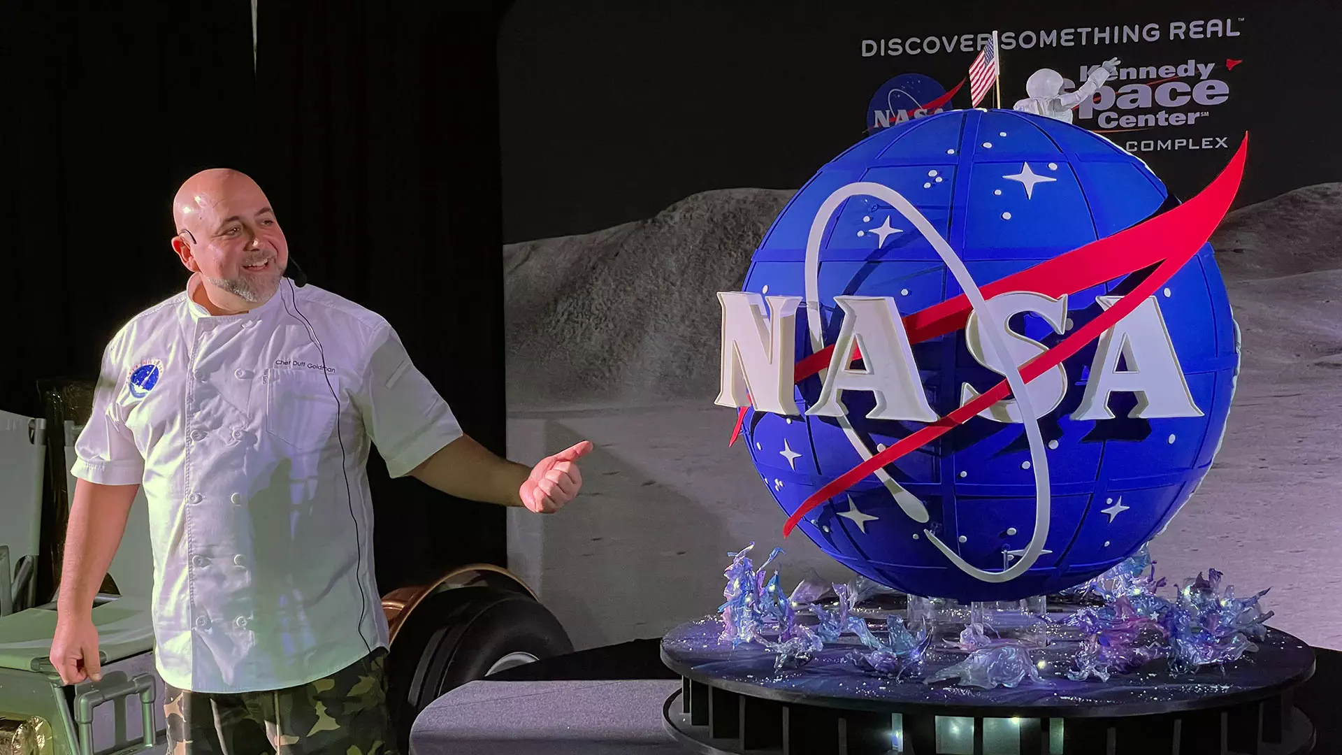 NASA Space Technology a man in a white t-shirt stands next to a large spherical cake that looks like nasa's famous 