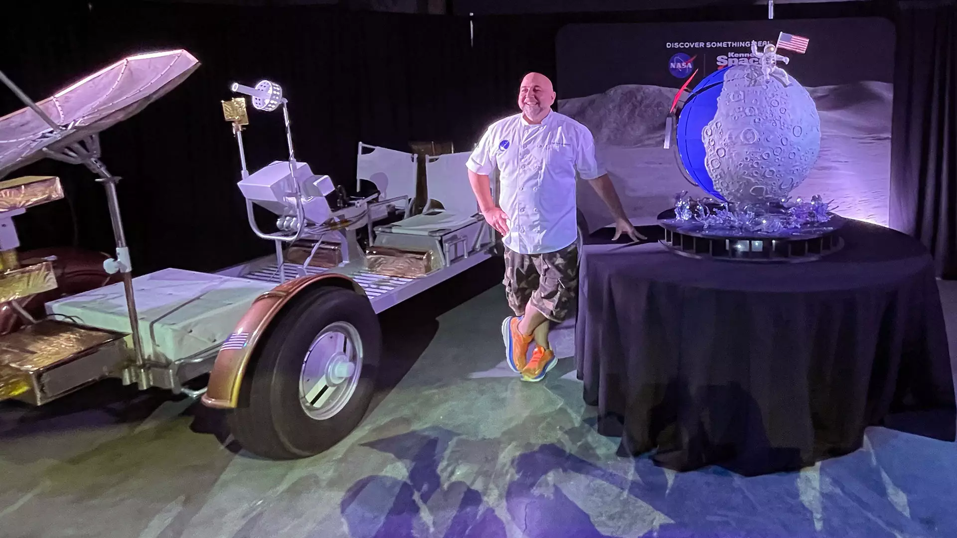 NASA Space Technology a smiling man in a white t shirt stands on a set next to a fake moon buggy and a cake that looks like the moon