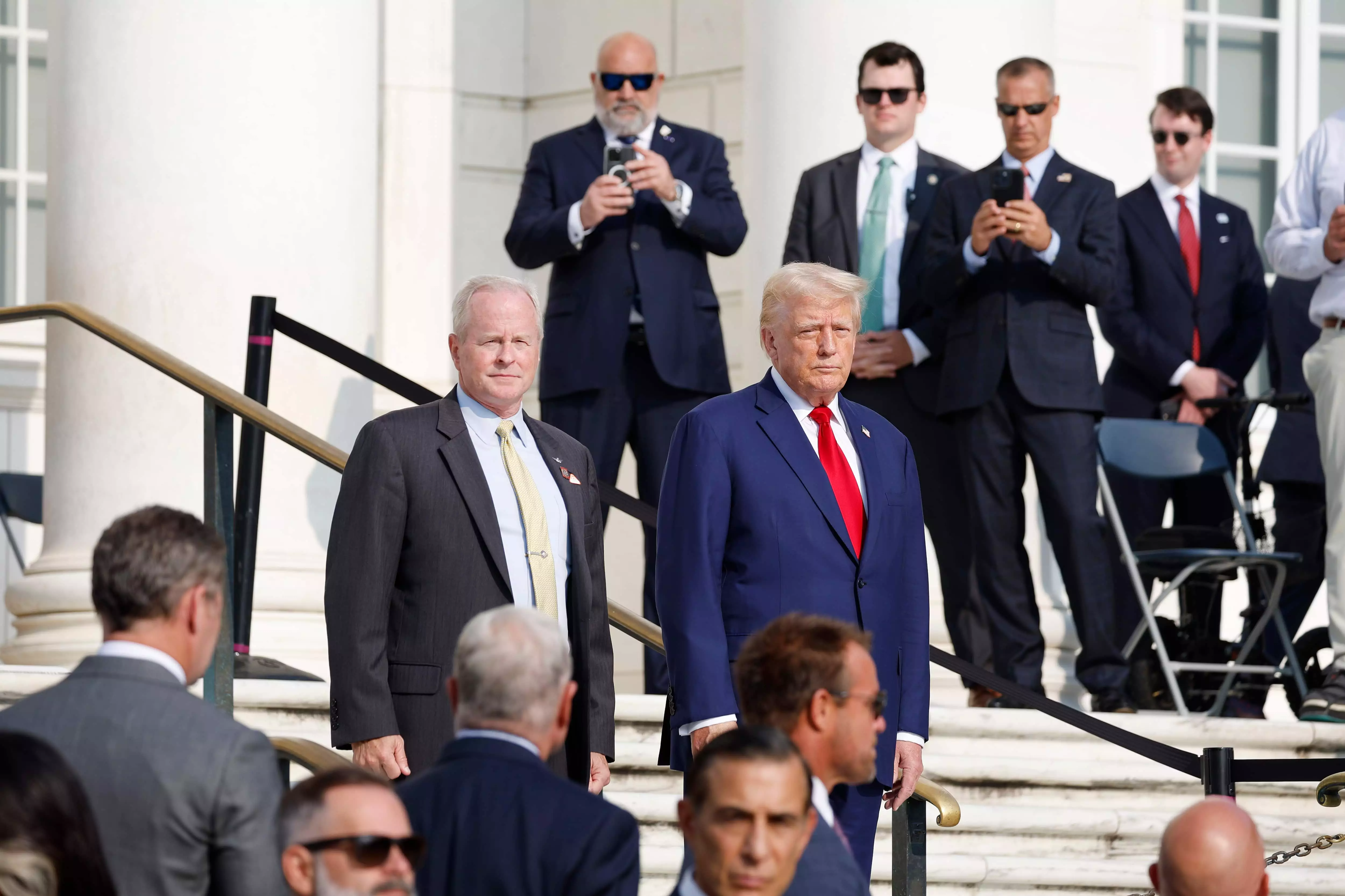 Politics tamfitronics Donald Trump walking down a flight of stairs, surrounded by men in suits.