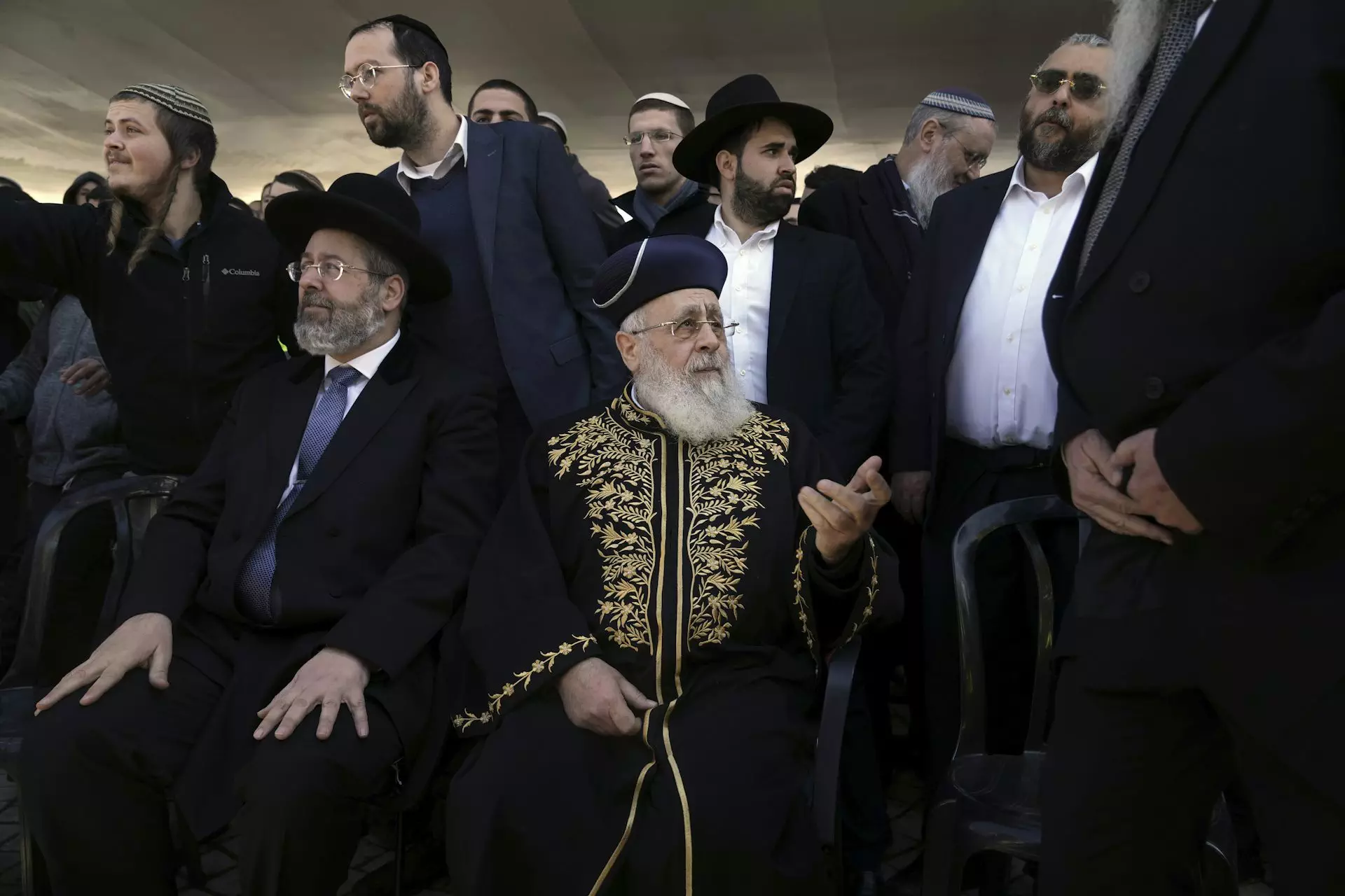 Politics tamfitronics A crowd of men wearing head coverings, with one man seated in front wearing an ornate gold and black robe.